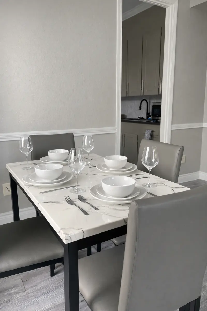 A neatly arranged dining table with white dishware, wine glasses, cutlery, and grey chairs, set against a light grey wall with a glimpse of a kitchen in the background.