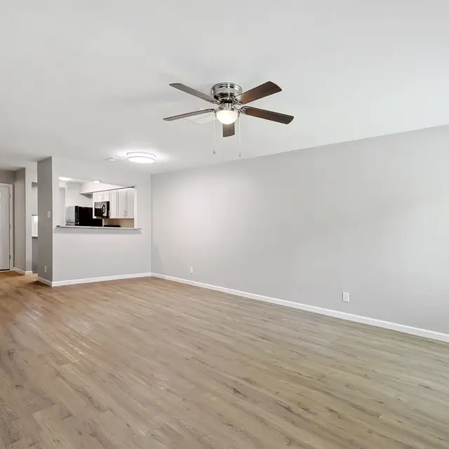 A spacious living room featuring light wooden flooring, a ceiling fan, and neutral-colored walls. There is a doorway leading to the kitchen area on the left side and a large empty wall on the right.