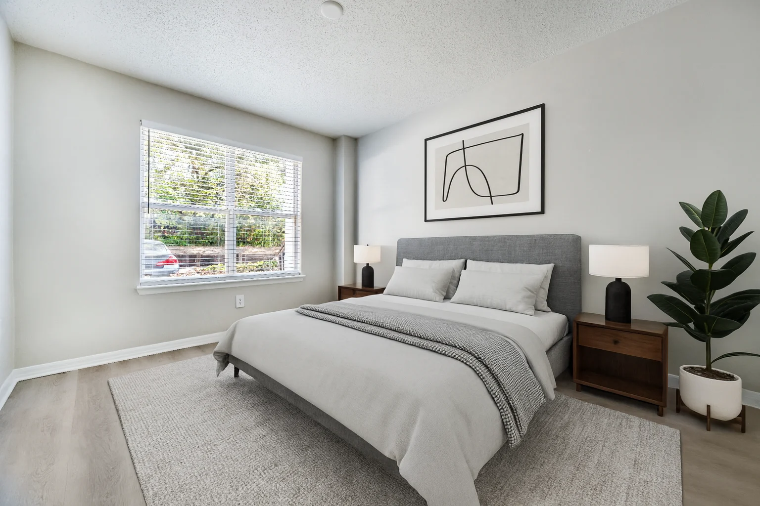 A modern bedroom featuring a gray bed with white bedding, two bedside lamps, a large window with blinds, and a green plant in the corner.