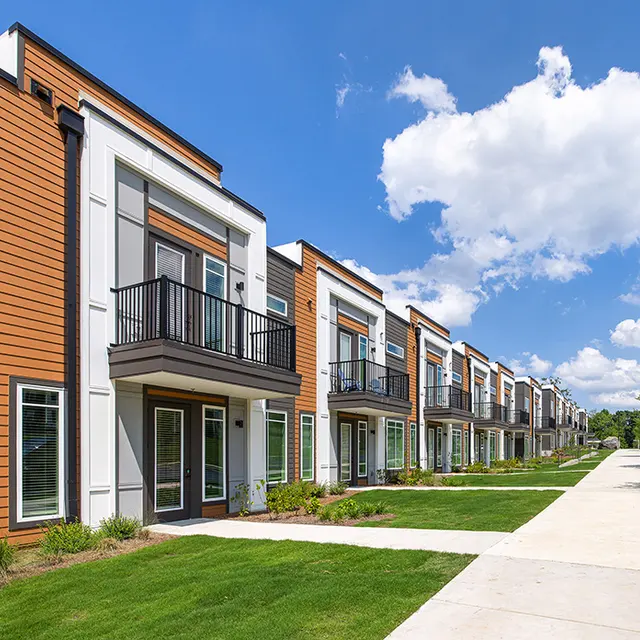 A row of modern townhomes situated along a paved walkway, featuring large windows and balconies, surrounded by green grass and trees under a blue sky with fluffy clouds.