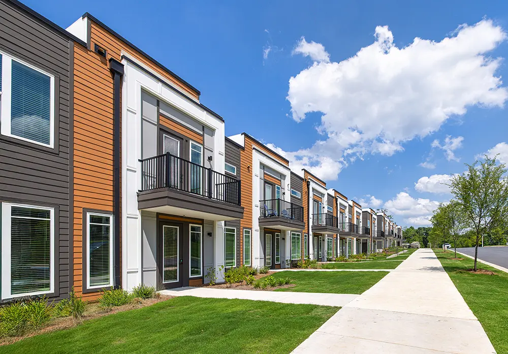 A row of modern townhomes situated along a paved walkway, featuring large windows and balconies, surrounded by green grass and trees under a blue sky with fluffy clouds.