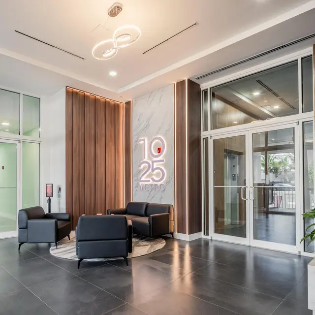 Modern lobby area with two black chairs, a decorative circular rug, and a plant, featuring a wall sign reading '10 25 METRO'.