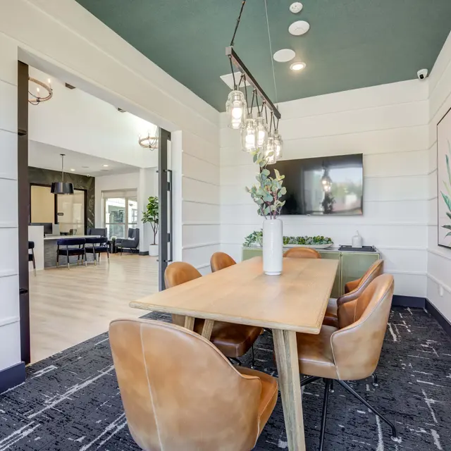 A modern meeting room featuring a wooden dining table surrounded by brown leather chairs, a decorative plant, and a modern light fixture. A large TV is mounted on the wall with artwork depicting a green plant. The room has a cozy atmosphere with stylish decor and a view into a larger lounge area.