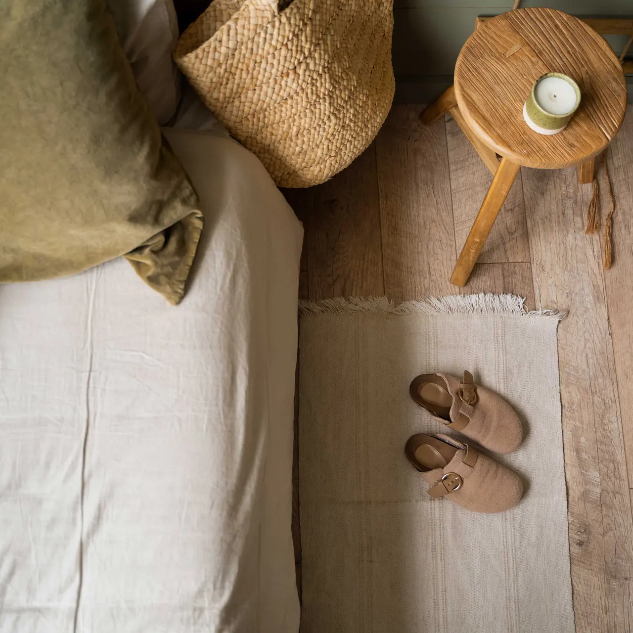 A cozy bedroom featuring a neatly made bed with light-colored linens, green pillows, a woven basket, a wooden stool with a candle, and a pair of slippers on a soft rug.