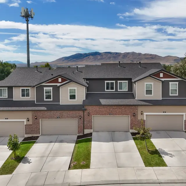 Aerial view of modern townhouses with brick facades and attached garages, set against a mountainous backdrop under a partly cloudy sky.