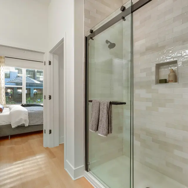 A modern bathroom with a glass shower enclosure, featuring beige tile walls and a towel hanging on a rack. In the background, a glimpse of a bedroom is visible through a doorway, showing a bed with patterned bedding and natural light coming in through the window.