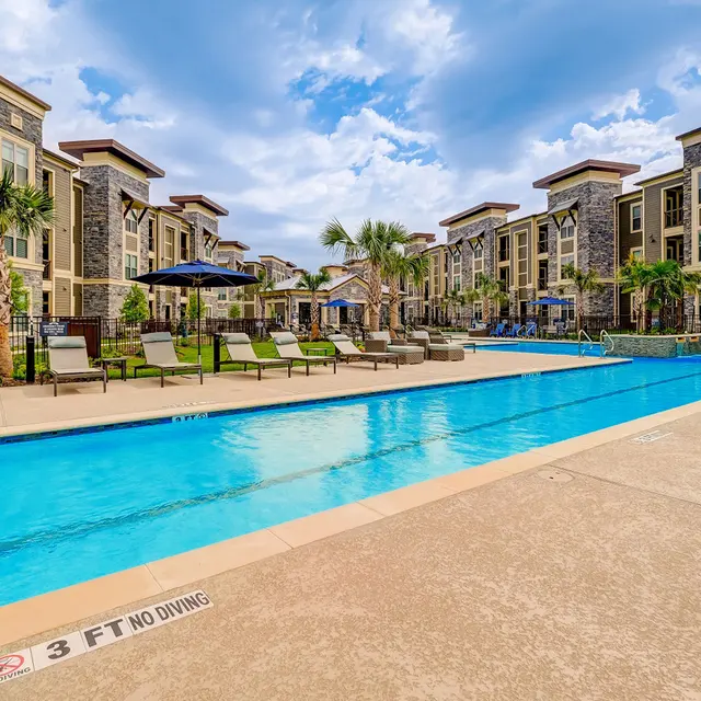 A spacious luxury apartment complex featuring a large pool surrounded by lounge chairs and palm trees, under a partly cloudy sky.