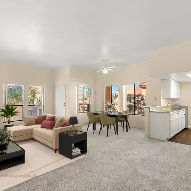 A spacious living room featuring a cozy seating area with a light-colored sofa, a black coffee table, and a gas fireplace. Large windows allow natural light to fill the room, complemented by a dining area with a round table. The kitchen is partially visible in the background, showing white cabinetry and modern appliances. The flooring is a mix of carpet and wood.