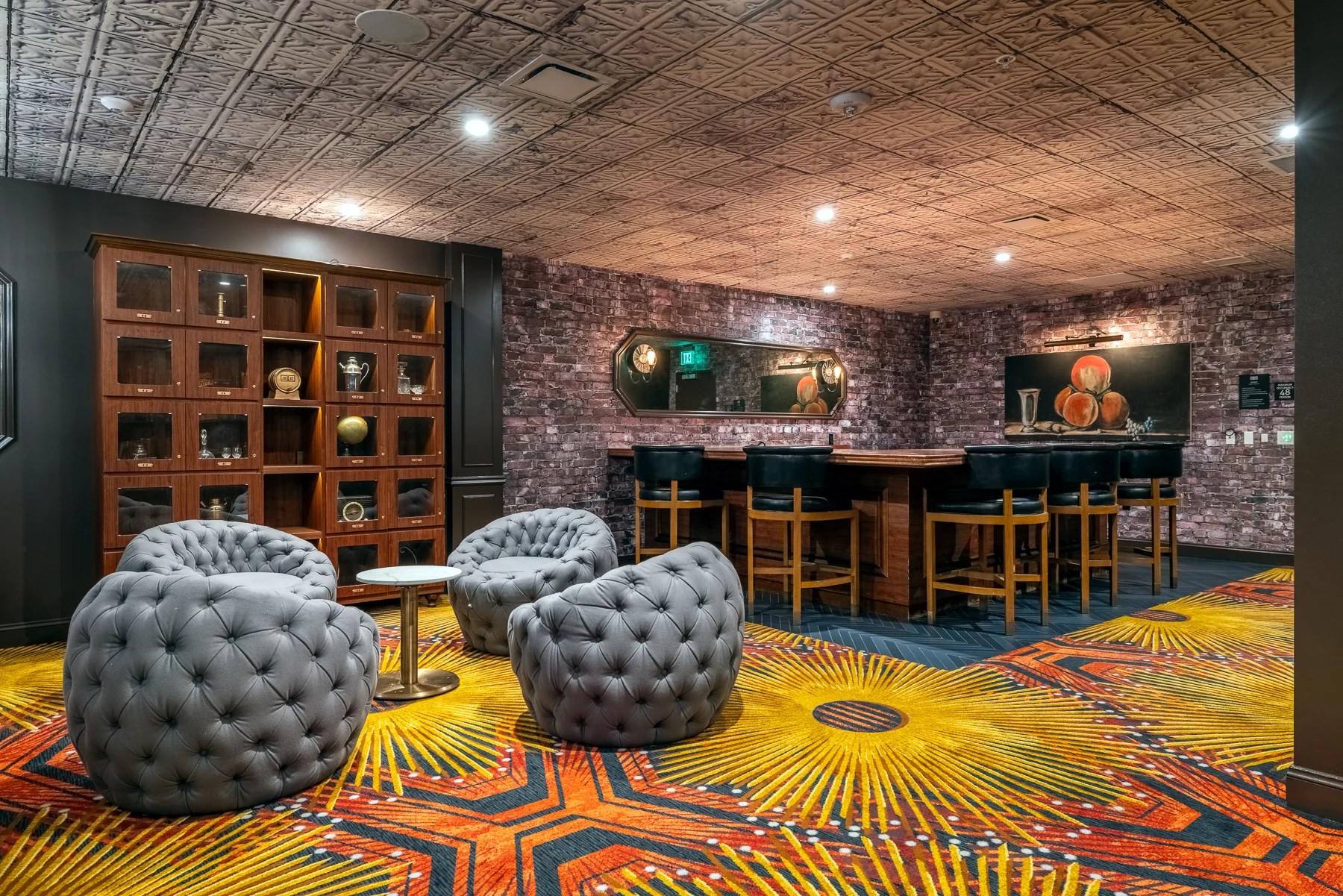 Modern Lounge Space A modern lounge area featuring plush tufted chairs, a circular coffee table, and a bar with high stools. The floor has a vibrant patterned carpet, and the walls are styled with exposed brick and dark wood shelving.