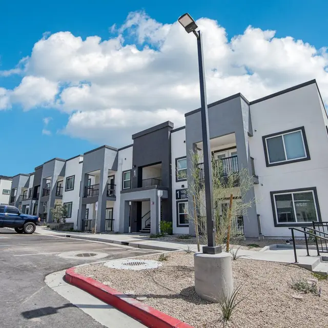 A modern apartment complex with multiple units, featuring a mix of white and gray exteriors, surrounded by desert landscape under a blue sky with clouds.