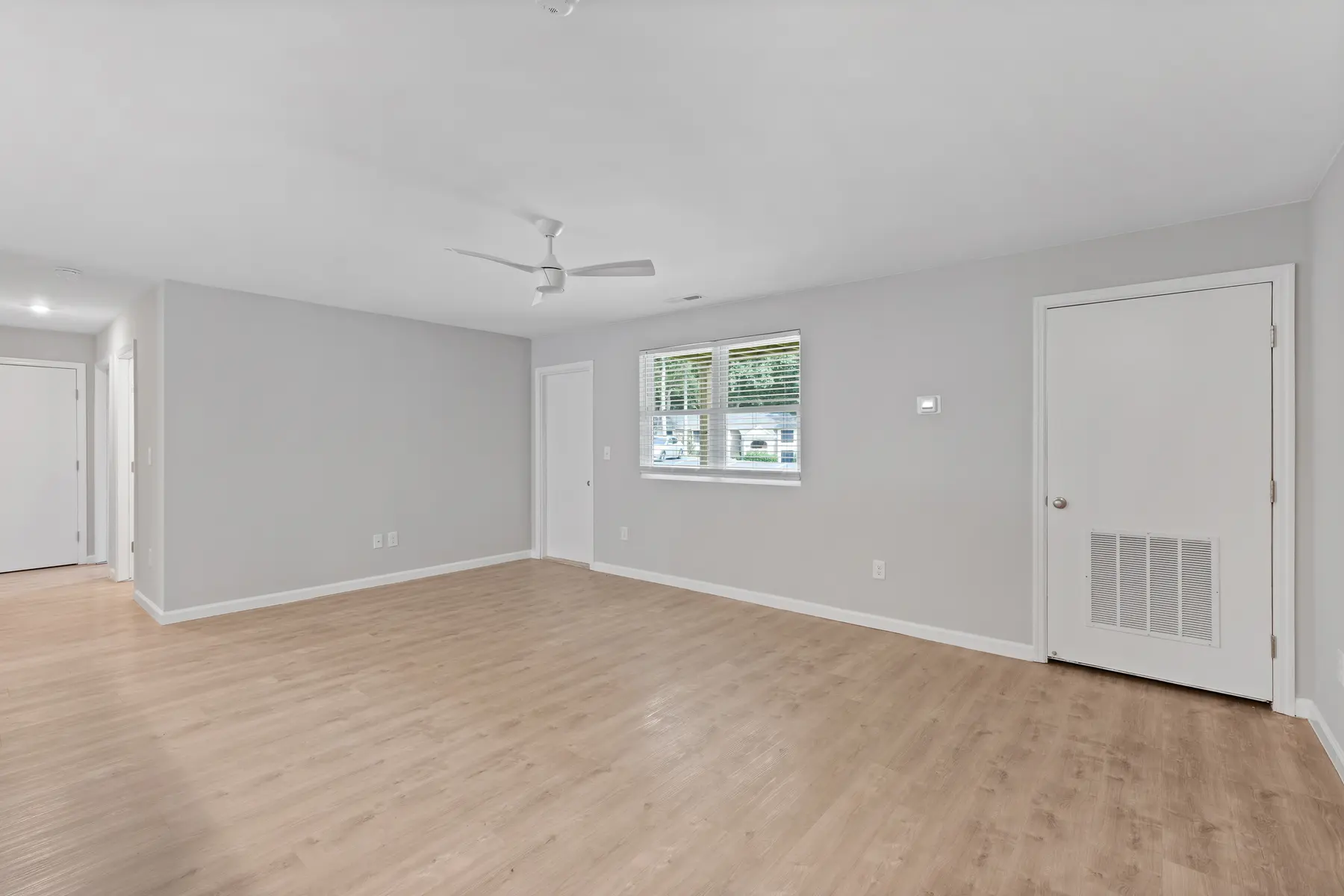 A spacious, empty room featuring light wood flooring, a ceiling fan, and a window with natural light. It has neutral gray walls and a door leading to another area.