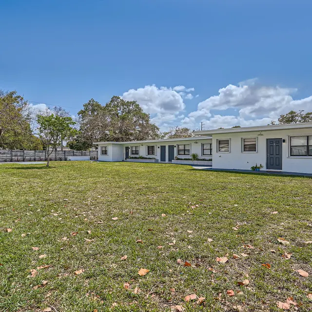 A spacious exterior view of a white house surrounded by lush green grass and trees under a clear blue sky with a few clouds.