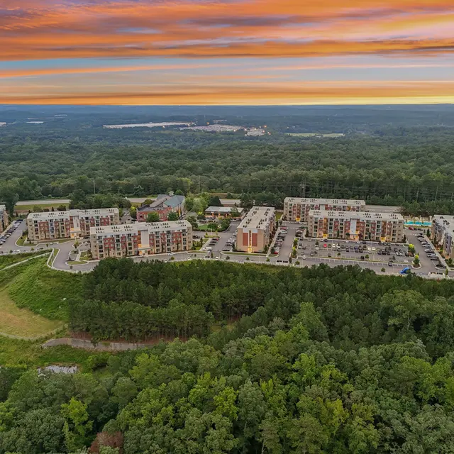 Aerial view of a modern apartment complex surrounded by lush greenery and forests, with a colorful sunset sky overhead.