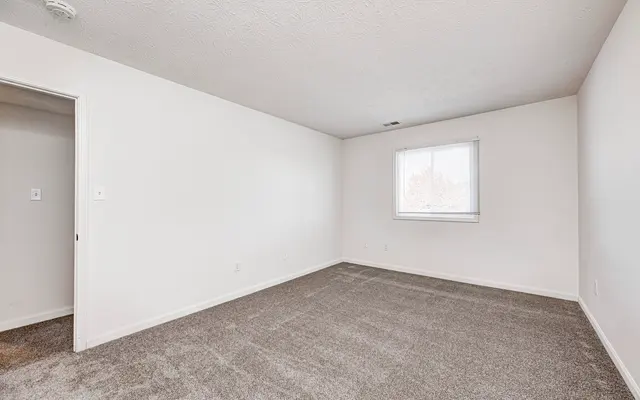 An empty room with light gray carpet and white walls. A single window on one wall lets in natural light.