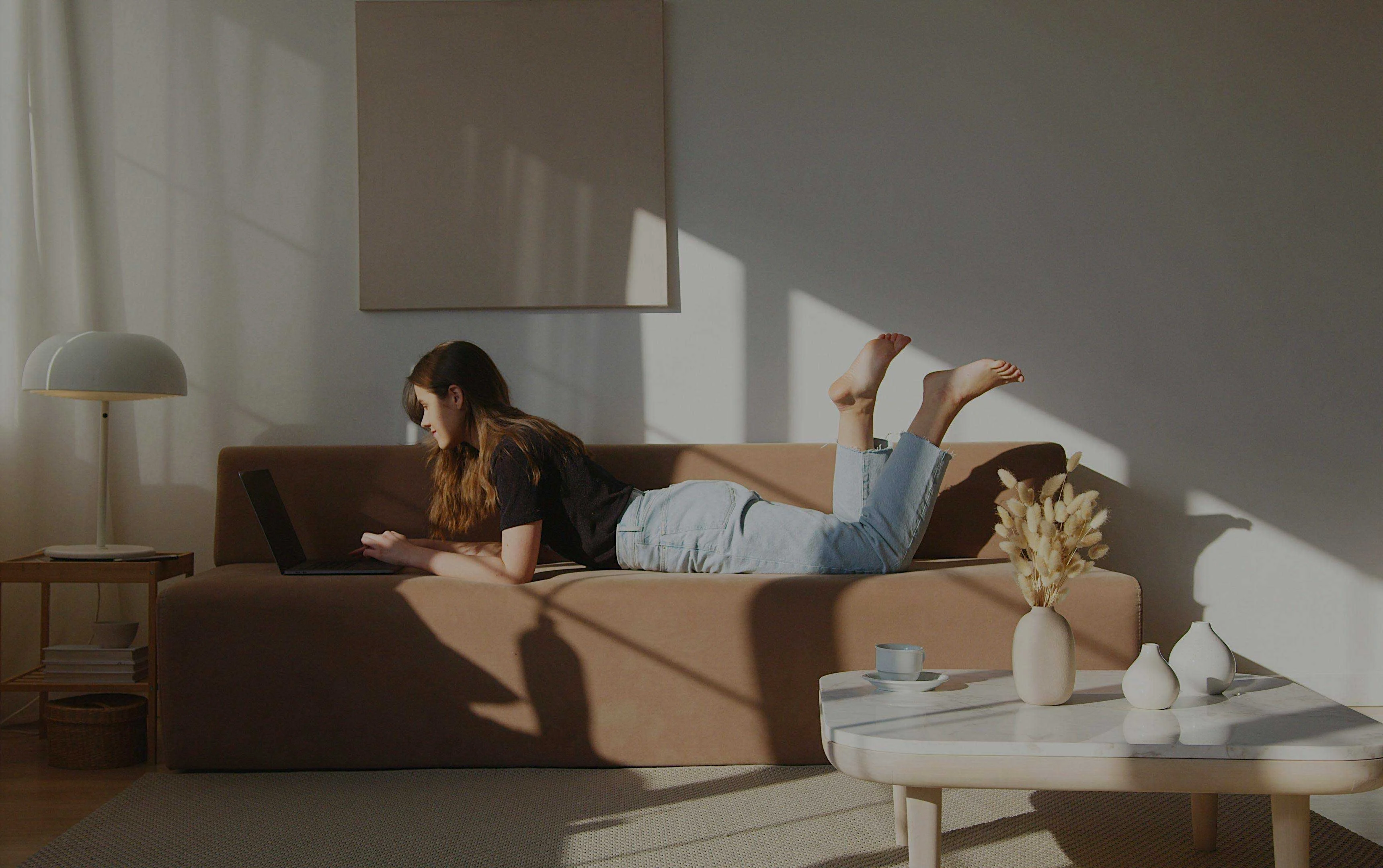 A young woman lying on a brown couch with her feet in the air, using a laptop. She is in a well-lit room with shadows and sunlight filtering through the curtains. A small table nearby holds a cup and decorative items.