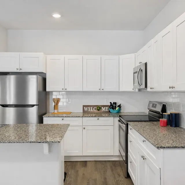 A modern kitchen featuring white cabinetry, granite countertops, stainless steel appliances, and a welcoming sign on the countertop.
