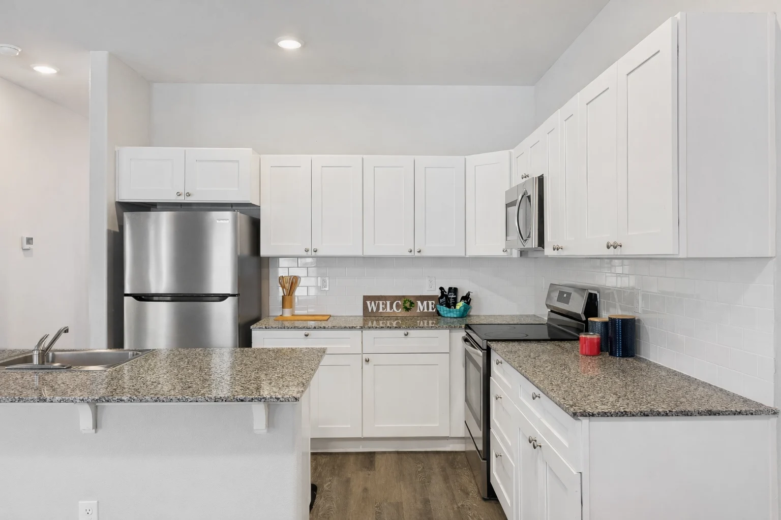 A modern kitchen featuring white cabinetry, granite countertops, stainless steel appliances, and a welcoming sign on the countertop.