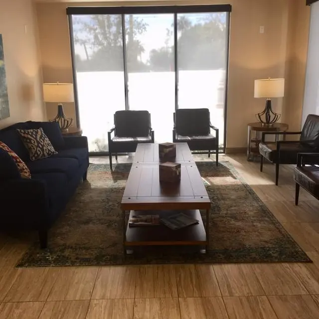A modern living room featuring a dark blue sofa, two black armchairs, a wooden coffee table, and two lamps. The room has large windows allowing natural light to come in, with a painting on the wall and a patterned rug on the floor.