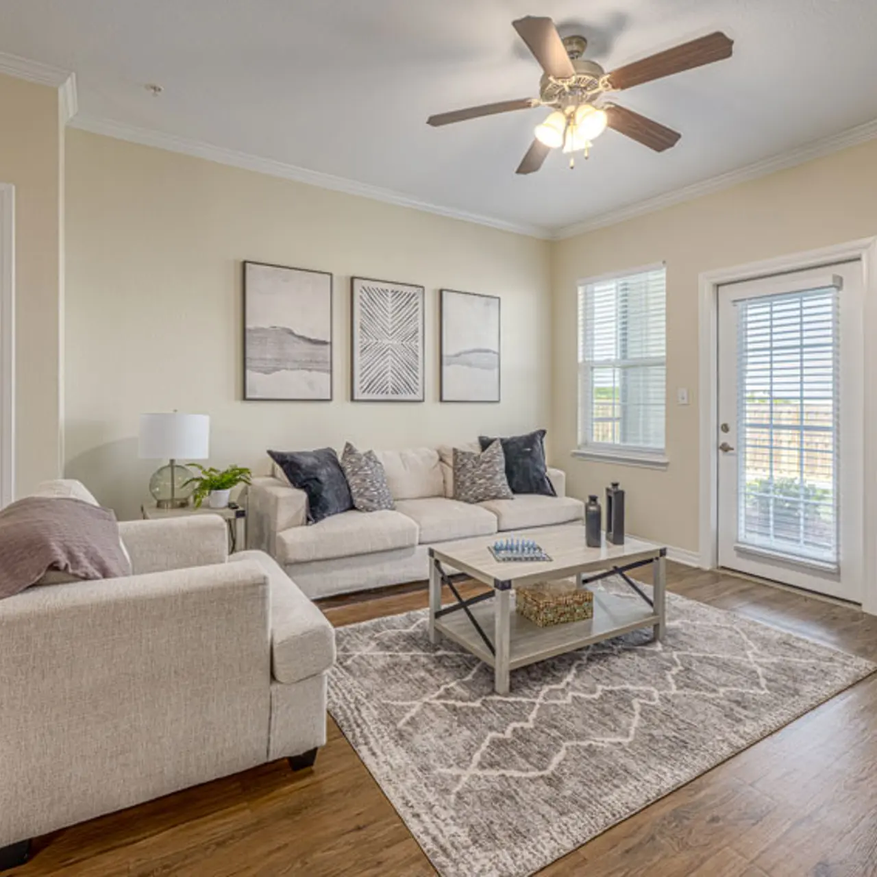A cozy living room featuring a beige sectional sofa and armchair, a coffee table in the center, and large windows allowing natural light. There are framed artworks on the wall and a door leading to a patio.