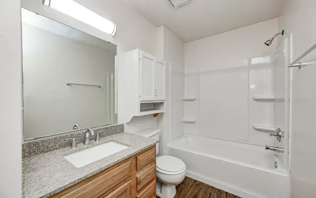 A modern bathroom featuring a light-colored wall, a wooden vanity with a granite countertop, a large mirror, and a tub-shower combo. The flooring is wooden, and the space is well-lit.