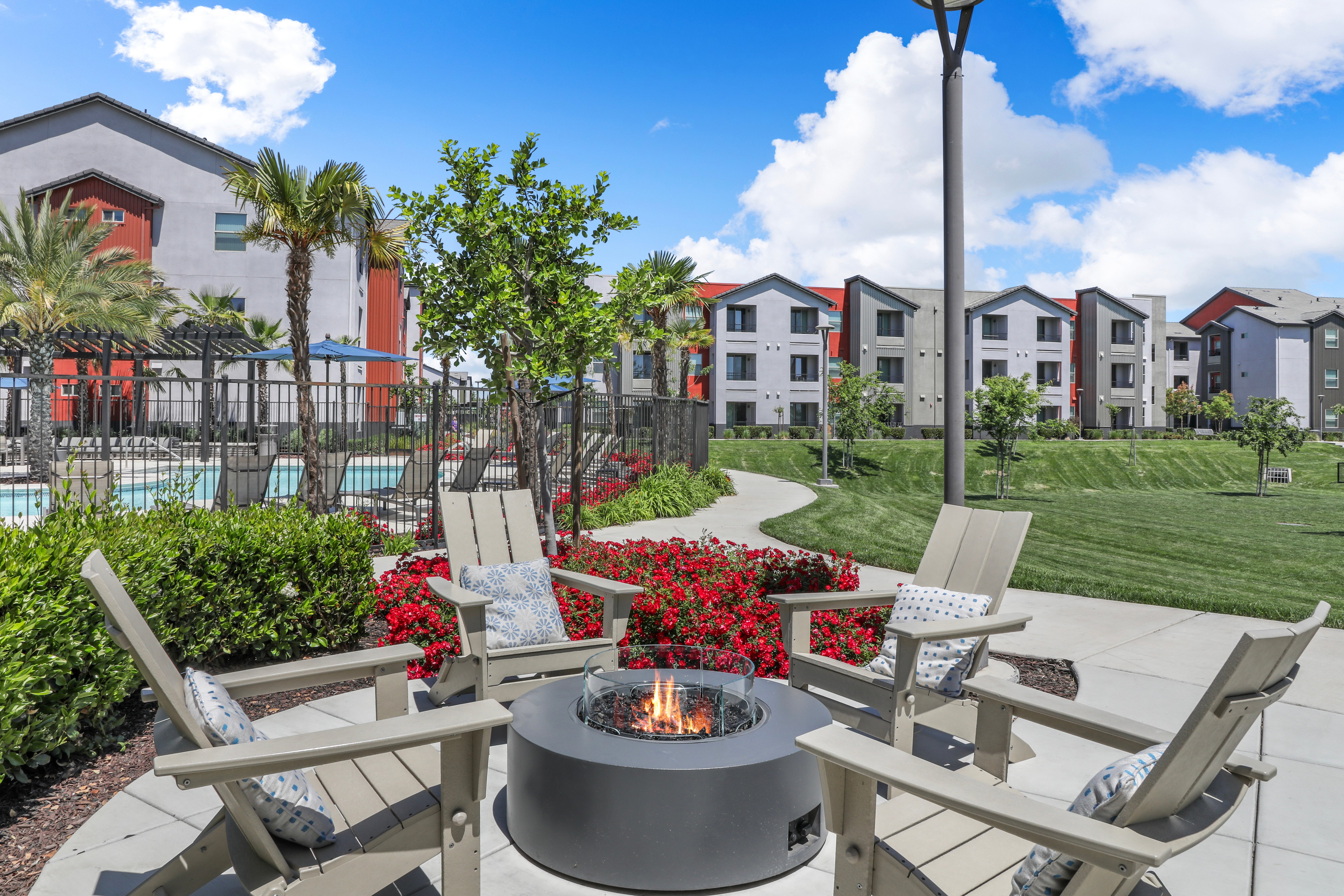 A cozy outdoor space featuring four lounge chairs surrounding a modern fire pit. In the background, a well-maintained lawn and apartment buildings are visible under a bright blue sky.