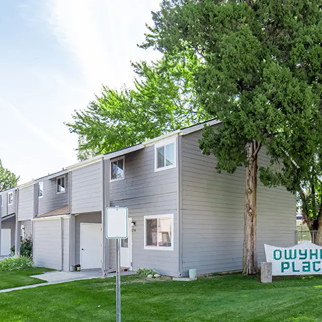 Owyhee Place Residential Complex A gray multi-unit residential building with a sign reading 'Owyhee Place' in front, surrounded by green grass and trees under a clear sky.