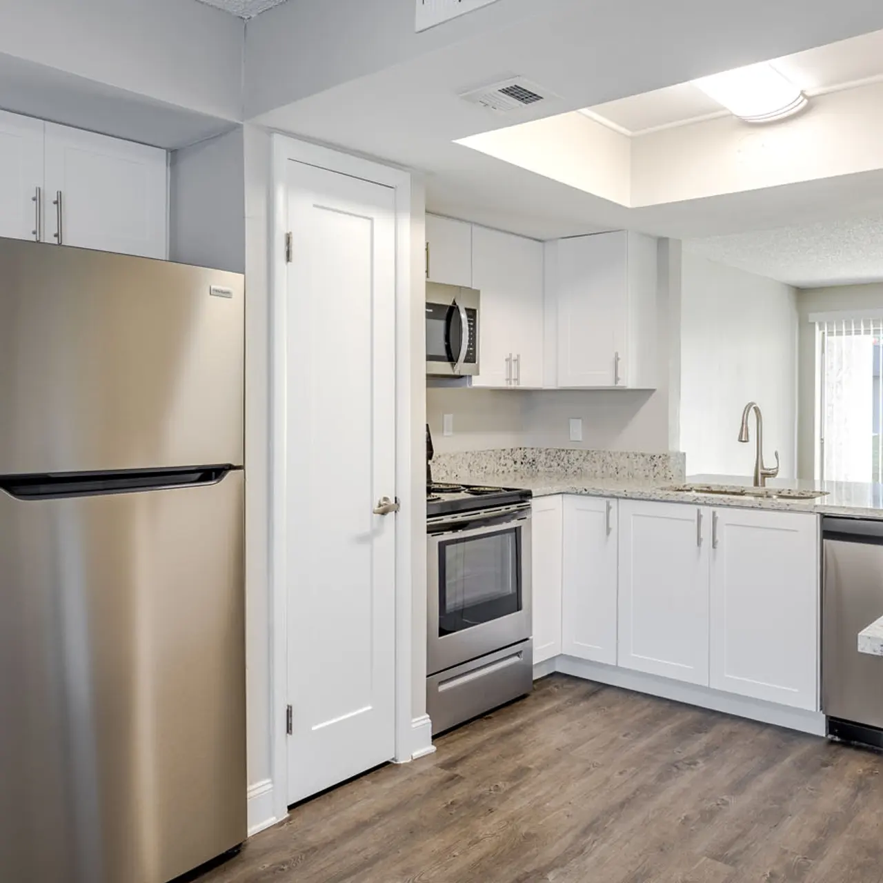 A modern kitchen with stainless steel appliances, including a refrigerator and stove, granite countertops, and white cabinetry. The space features a large window letting in natural light, and a wooden floor adds warmth to the design.