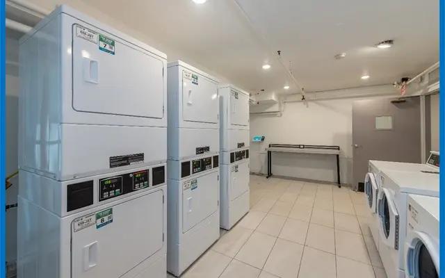 A clean laundry room featuring stacked washing and drying machines along one wall and additional dryers on the side. The floor is tiled and well-lit, with a folding station against the wall and a door in the background.