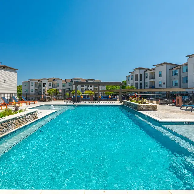 A large swimming pool surrounded by sun loungers in a well-maintained apartment complex under a clear blue sky.