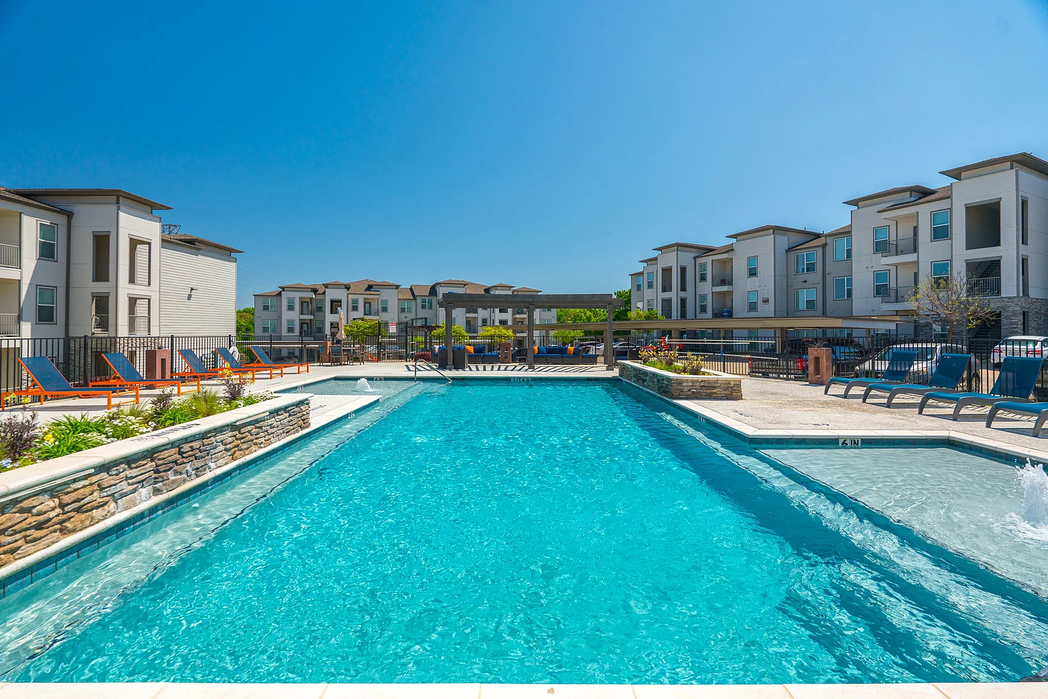 Apartment Complex Swimming Pool A large swimming pool surrounded by sun loungers in a well-maintained apartment complex under a clear blue sky.