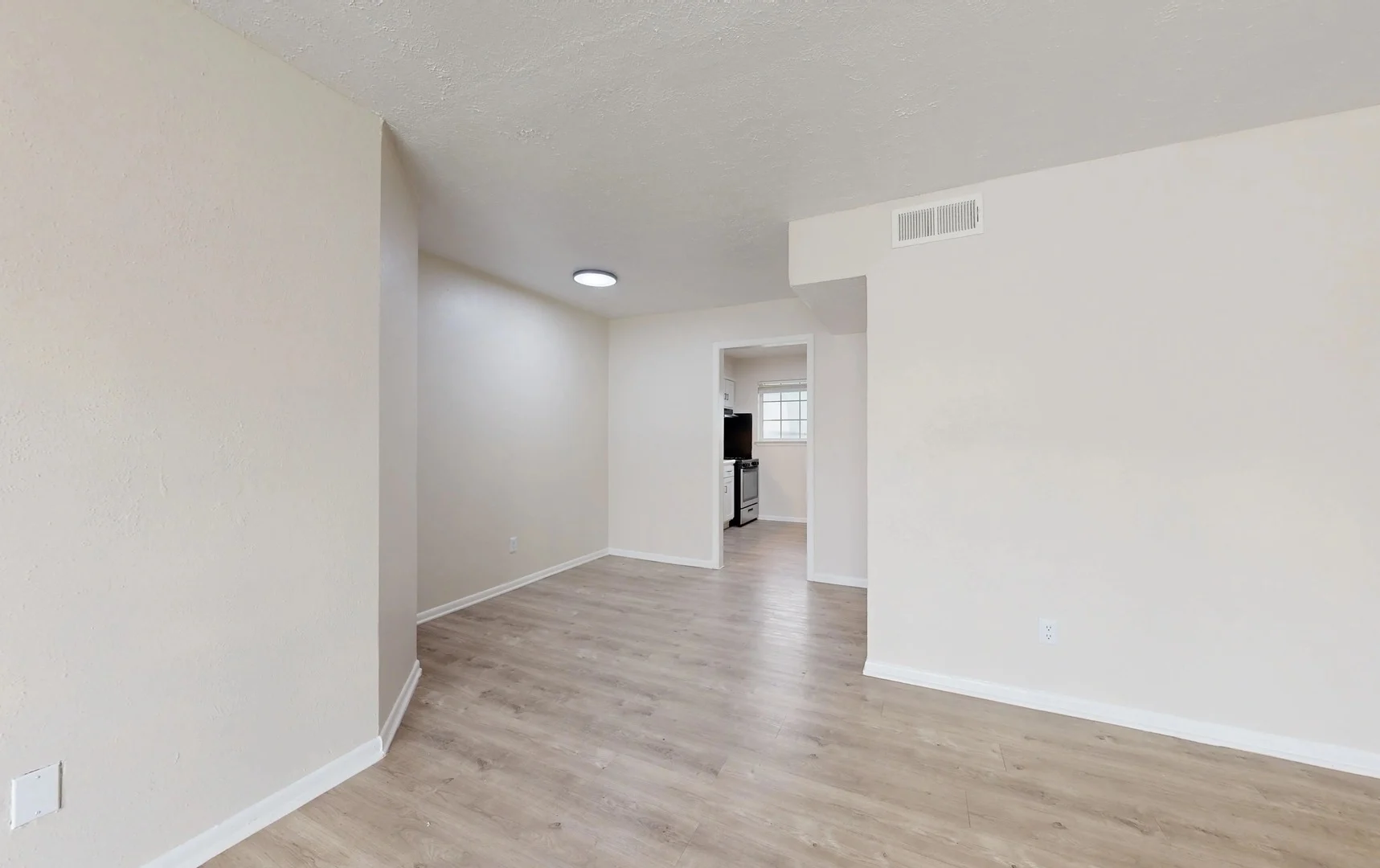 Empty Room Interior A spacious, empty room featuring light-colored walls and hardwood flooring. There is a doorway leading to another room visible in the background.