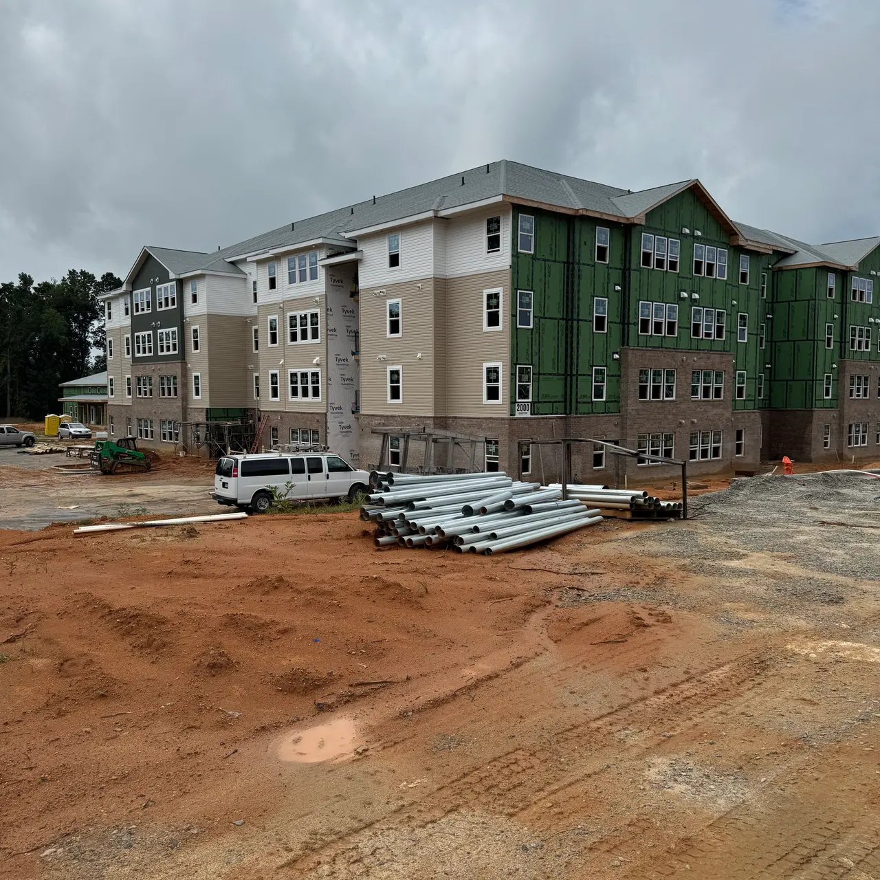 A construction site featuring a partially finished multi-story building with beige and green exterior. Piles of pipes are visible in the foreground, along with heavy machinery and excavated soil.