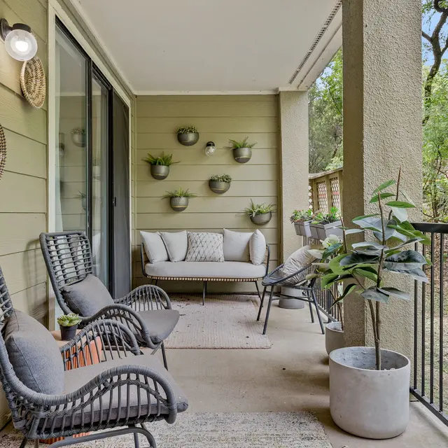 A stylish balcony with two wicker chairs and a sofa, surrounded by green foliage and a railing. Wall-mounted planters display small plants.
