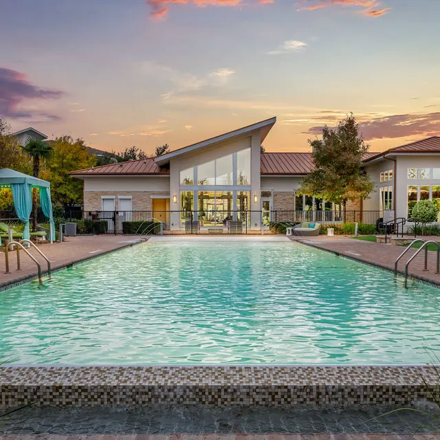 A scenic swimming pool at sunset, featuring turquoise cabanas, green lounge chairs, and a modern building in the background.