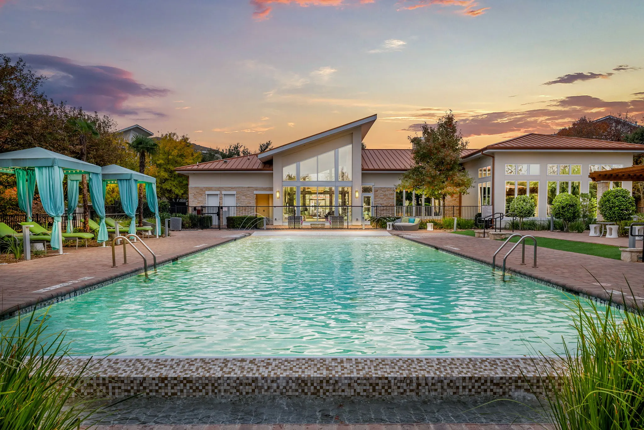 A scenic swimming pool at sunset, featuring turquoise cabanas, green lounge chairs, and a modern building in the background.