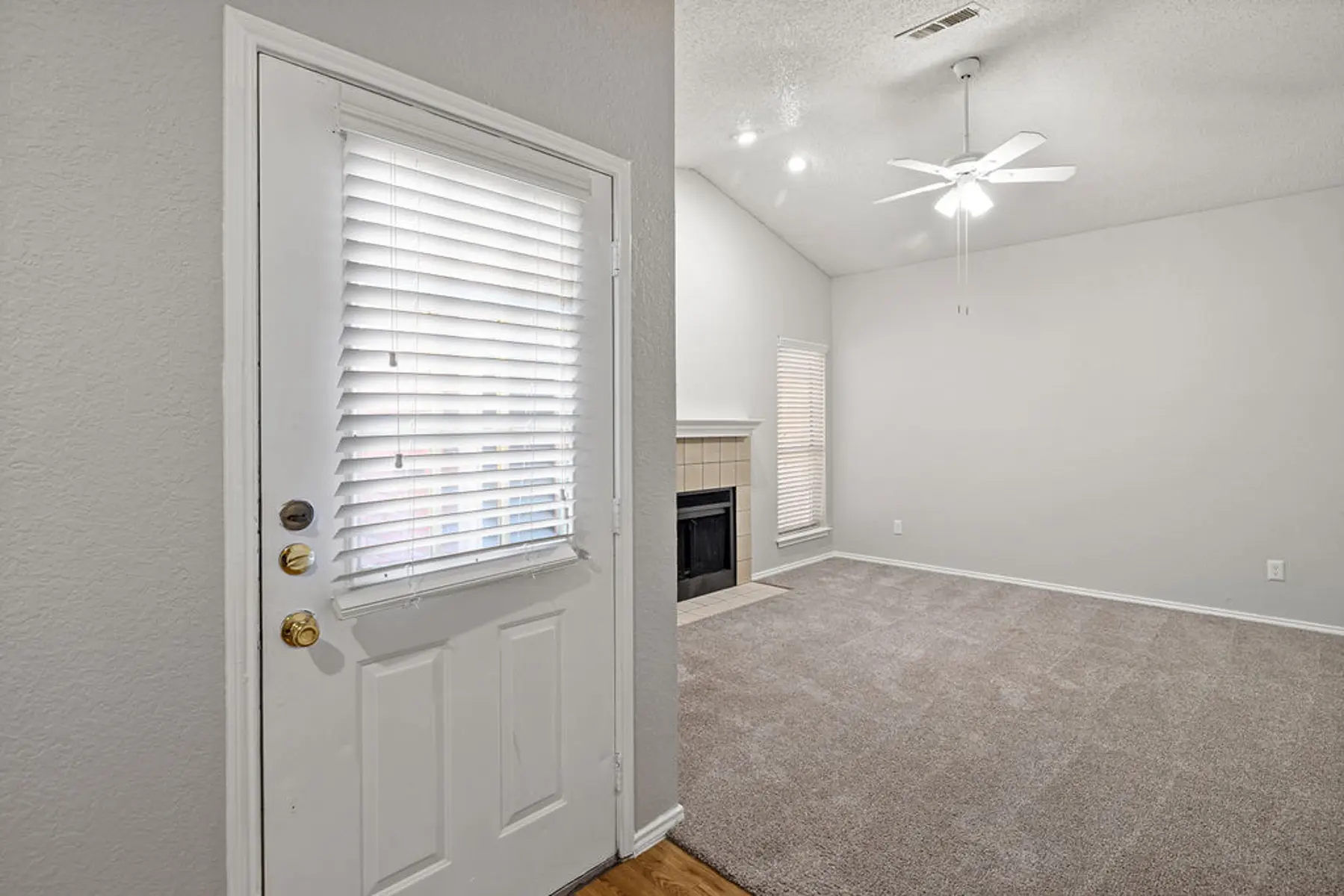 Cozy Living Room Entry Interior view of a living room with an entrance door on the left, carpeted floor, and a ceiling fan. The room features a fireplace and a window with blinds.