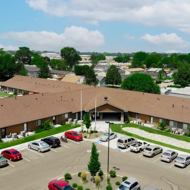 Aerial View of a Residential Complex Aerial view of a residential complex featuring multiple buildings with brown roofs, surrounded by neatly maintained green lawns and landscaped areas. A parking lot with several cars is visible in the foreground, along with trees and other vegetation.
