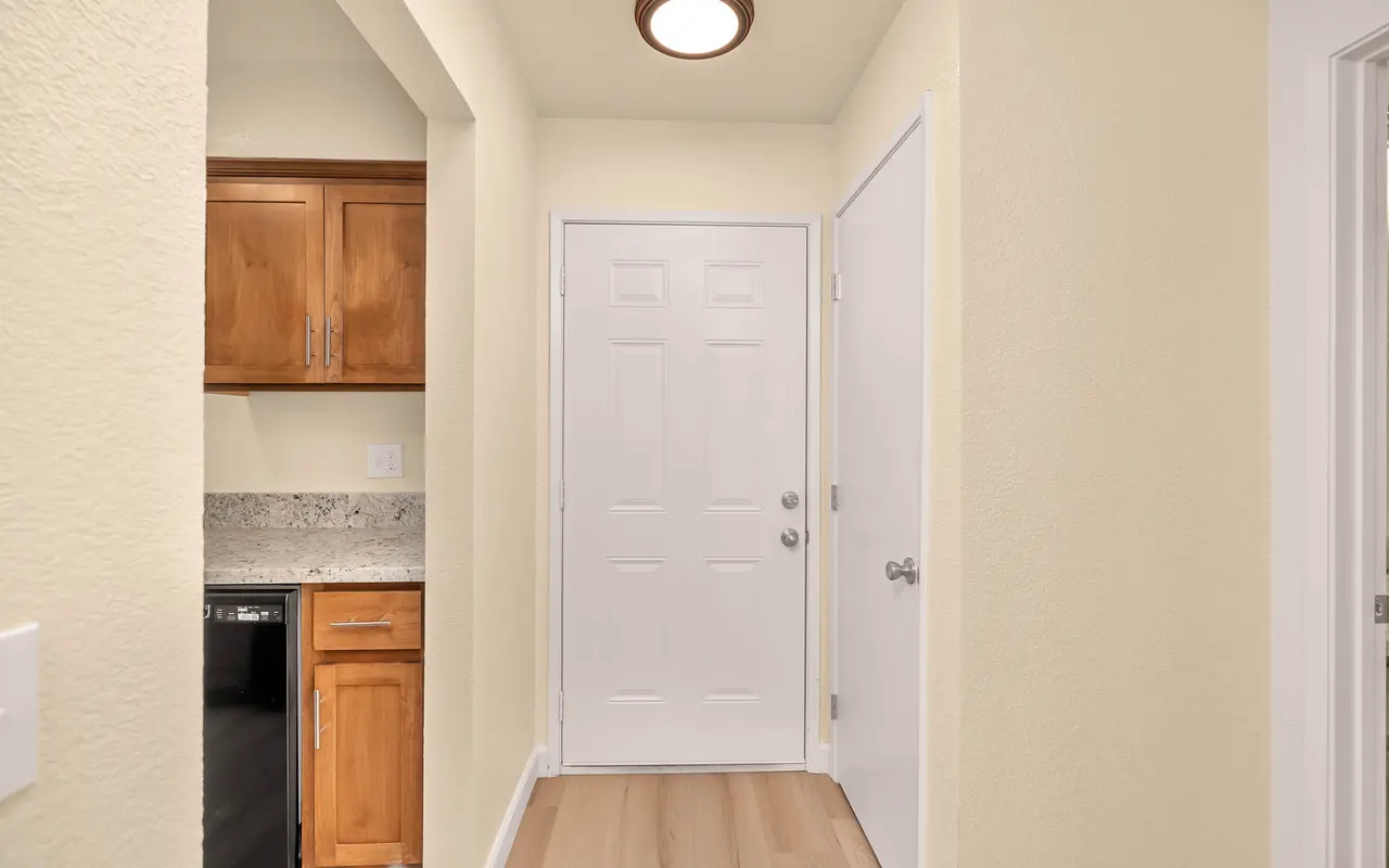 View of a hallway leading to a white door with a light fixture overhead. To the left, there are wooden cabinets and a countertop. The floor appears to be light-colored wood.