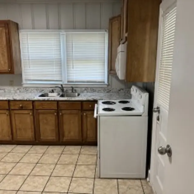 A kitchen with wooden cabinets, a white stove, and a sink beneath a window with blinds. The floor is tiled, and a door is visible on the right side.
