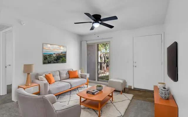A bright and modern living room featuring a light gray sofa with orange cushions, a small orange coffee table, and a chair. A wall-mounted TV is present, along with a lamp and artwork depicting a landscape. The room has a sliding glass door leading outside, with natural light coming in.
