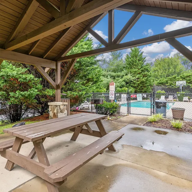 A wooden pavilion with a picnic table overlooking a swimming pool area. The pavilion has a wooden roof and is surrounded by greenery. In the background, there are lounge chairs by the pool, a fence, and a clear blue sky with a few clouds.