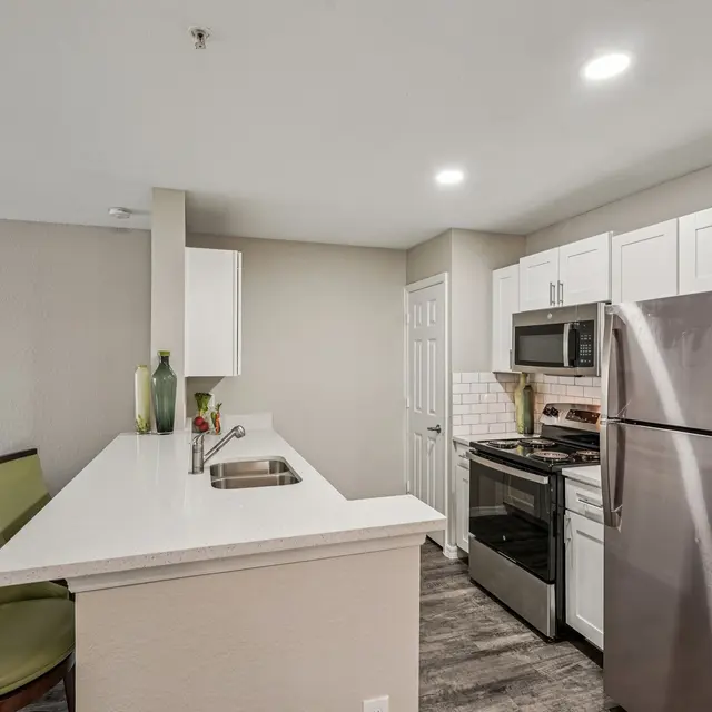 A modern kitchen featuring white cabinets, stainless steel appliances, and a sleek countertop. The kitchen island has green bar stools, and there's a hint of a living area in the background.