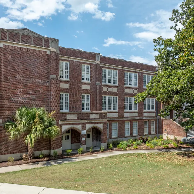 A historic red brick school building with large windows and multiple stories surrounded by greenery and palm trees.