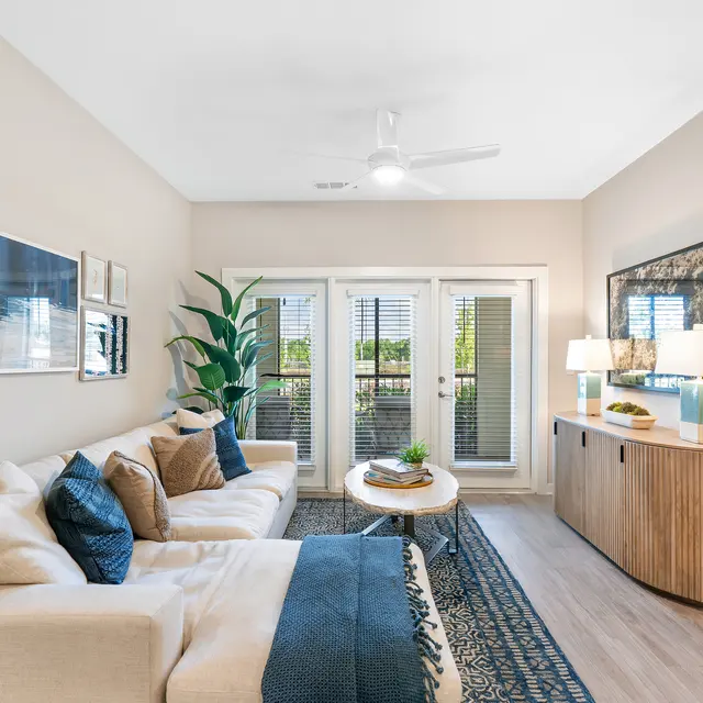 Chic Modern Living Room A modern living room featuring a light-colored sofa with blue and white pillows, a wooden side table with lamps, and large windows with vertical blinds. A plant is present in one corner, and a patterned area rug covers the hardwood floor.
