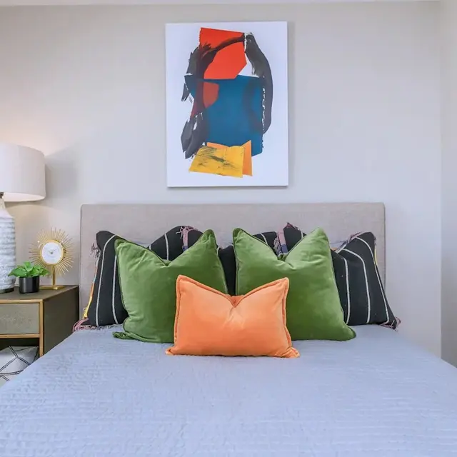 Cozy bedroom featuring a bed with colorful pillows, a bedside table, a lamp, and an abstract painting on the wall.