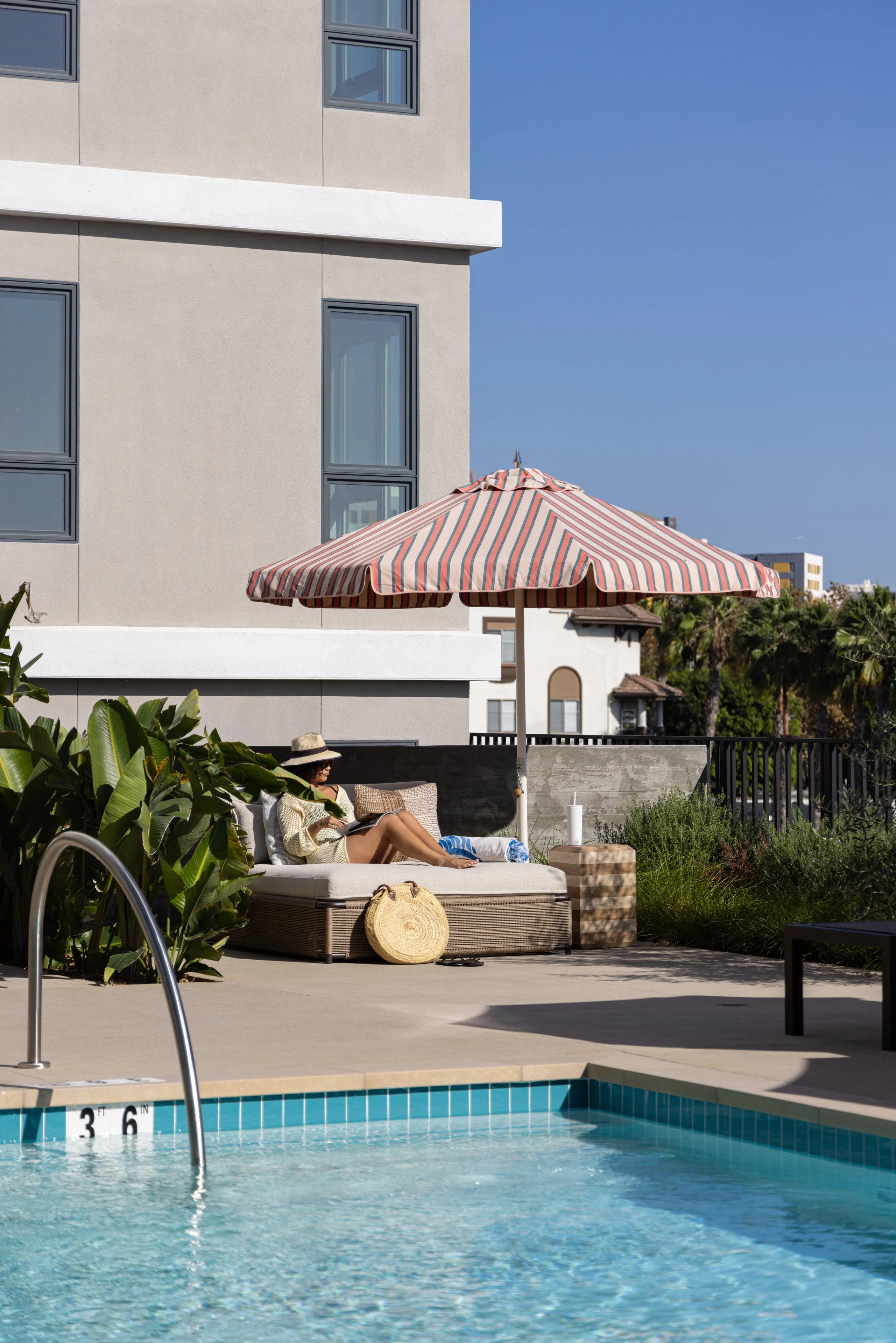 Sunny Poolside Retreat A tranquil poolside scene featuring a lounging area under a striped umbrella, with a clear blue pool in the foreground and modern apartment building in the background.