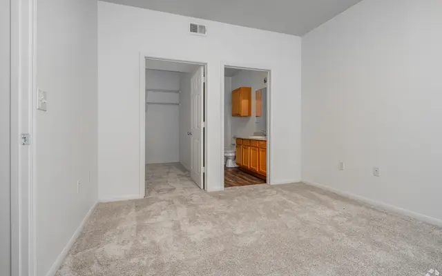 A vacant room featuring carpet, white walls, and two doorways leading to other spaces. One doorway leads to a closet, while the other opens to a kitchen area with wooden cabinets. The room is well-lit and spacious.