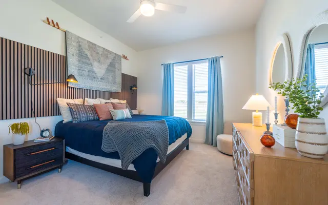 A modern bedroom featuring a large bed with blue bedding and various decorative pillows. The headboard is designed with wooden slats and a large wall art above it. There’s a nightstand on the left with a plant, and a dresser on the right. The room has two windows with light curtains, a small stool, and a warm color palette.
