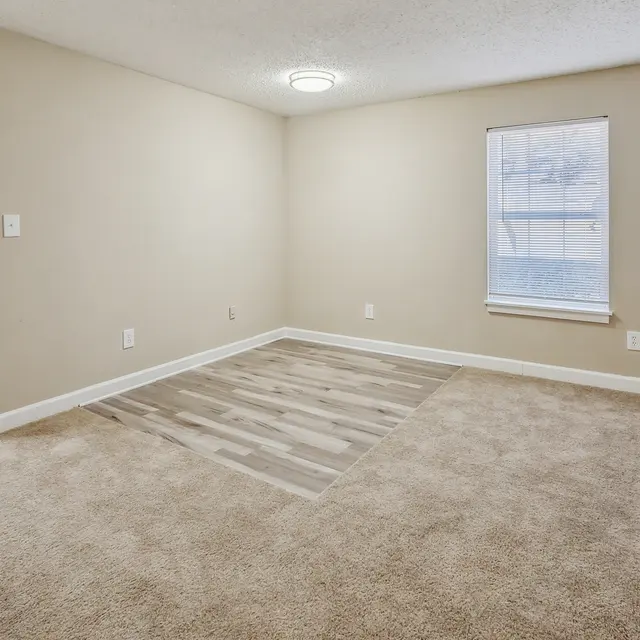 A spacious, empty living room with beige walls and carpet. Two windows allow natural light to enter, and a section of the floor has different wood-like flooring. A door leads to another room.