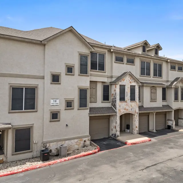 A three-story apartment building featuring light-colored walls and stone accents, with a red parking area in front and green trees in the background.